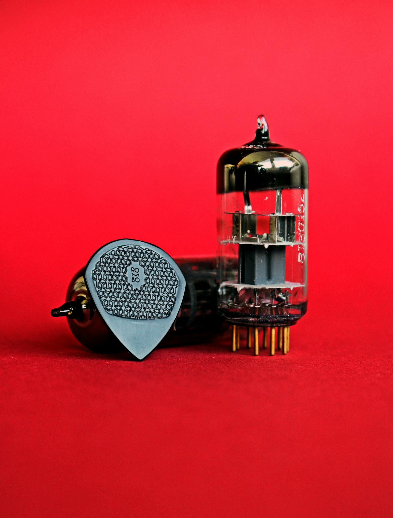 Close-up of a guitar pick and vacuum tube on a vibrant red background.